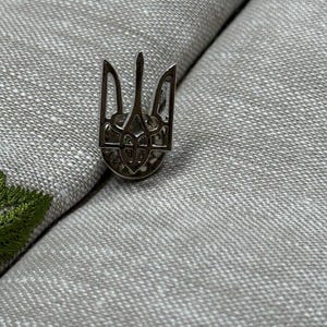 Könnte beinhalten: Eine silberfarbene Anstecknadel mit dem Tryzub, dem Nationalemblem der Ukraine. Die Nadel hat ein detailliertes, dreizackiges Trident-Design, vor einem strukturierten, hellgrauen Hintergrund. Die Nadel ist ein kleines, dekoratives Accessoire.