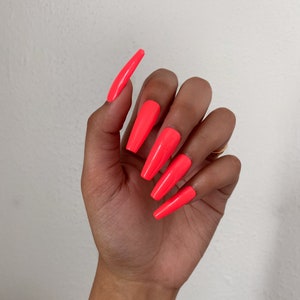 May include: A hand with long, bright neon-coral colored, coffin-shaped acrylic nails. The nails are uniformly shaped and have a glossy finish. The hand is positioned against a plain white background.