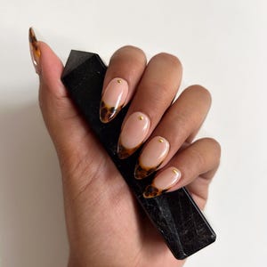 May include: Oval-shaped nails with a tortoiseshell design on the tips and small gold studs. The nails are a light pink color. A black, rectangular object is held in the hand.