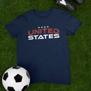Op de afbeelding: Een marineblauw t-shirt met een witte afbeelding waarop "UNITED STATES" in rode letters staat met vijf witte sterren boven het woord "UNITED".