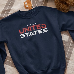 Op de afbeelding: Een donkerblauw sweatshirt met de tekst "UNITED STATES" in rood en wit. De tekst is gestyled met een vintage look.