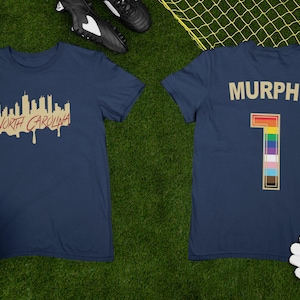 May include: A navy blue t-shirt with a gold skyline graphic and the text "NORTH CAROLINA" on the front. The back of the shirt has the name "MURPHY" and the number "1" with a rainbow design.