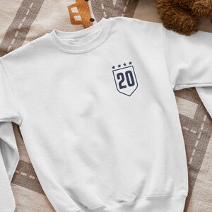K&ouml;nnte beinhalten: Wei&szlig;er Pullover mit einem marineblauen Patch mit der Nummer 20 und f&uuml;nf Sternen.