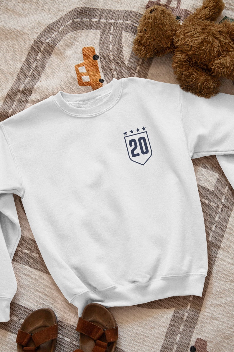 K&ouml;nnte beinhalten: Wei&szlig;er Pullover mit einem marineblauen Patch mit der Nummer 20 und f&uuml;nf Sternen.