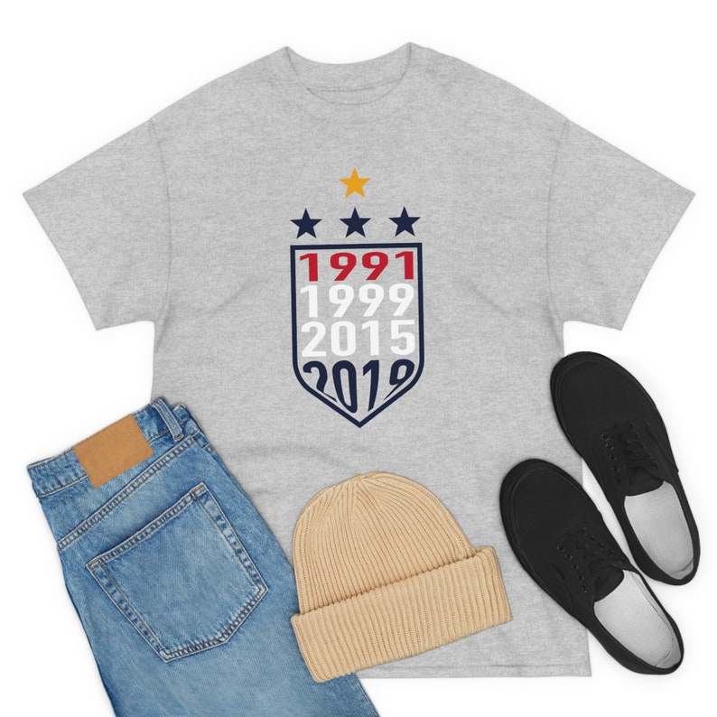 Puede incluir: Una camiseta gris con un dise&ntilde;o de escudo azul y rojo que presenta los a&ntilde;os 1991, 1999, 2015 y 2019. El escudo est&aacute; coronado con tres estrellas doradas.
