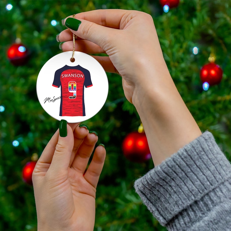 Op de afbeelding: Ronde, witte keramische kerstversiering met een rood en marineblauw voetbalshirtontwerp. Het shirt heeft de naam "Swanson" en het nummer "9". Een gouden draad is bevestigd om op te hangen.