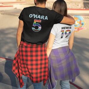 Puede incluir: Dos personas que llevan camisetas a juego de color negro y blanco con n&uacute;meros arco&iacute;ris en la espalda. La persona de la izquierda lleva una camisa de cuadros rojos y negros y la persona de la derecha lleva una camisa de cuadros morados y grises. El texto en la parte posterior de las camisetas dice "O'HARA 5" y "MEWIS 19".