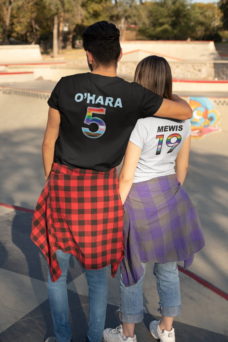 Puede incluir: Dos personas que llevan camisetas a juego de color negro y blanco con n&uacute;meros arco&iacute;ris en la espalda. La persona de la izquierda lleva una camisa de cuadros rojos y negros y la persona de la derecha lleva una camisa de cuadros morados y grises. El texto en la parte posterior de las camisetas dice "O'HARA 5" y "MEWIS 19".