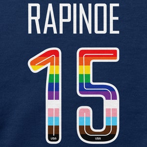 Puede incluir: Una camiseta azul con el nombre "RAPINOE" y el n&uacute;mero "15" en colores arco&iacute;ris con un contorno blanco. Los n&uacute;meros tienen un peque&ntilde;o logotipo "USA" en la parte inferior.