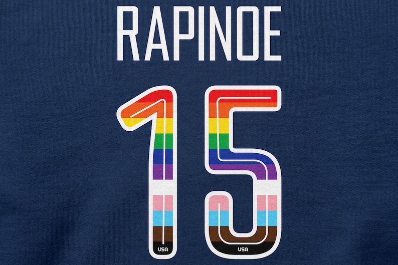 Puede incluir: Una camiseta azul con el nombre "RAPINOE" y el n&uacute;mero "15" en colores arco&iacute;ris con un contorno blanco. Los n&uacute;meros tienen un peque&ntilde;o logotipo "USA" en la parte inferior.