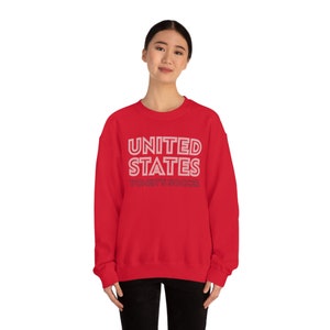 Puede incluir: Sudadera roja con el texto "UNITED STATES WOMEN'S SOCCER" en blanco con contorno rosa.