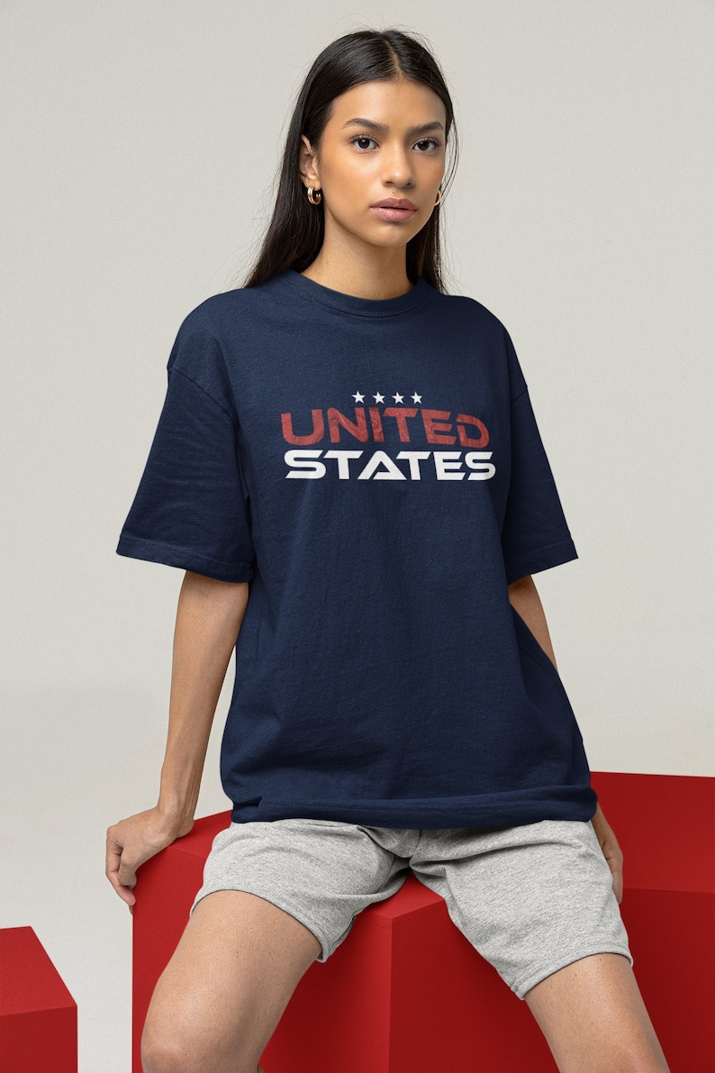 Op de afbeelding: Een marineblauw t-shirt met een witte afbeelding waarop "UNITED STATES" staat met vijf sterren boven het woord "UNITED".