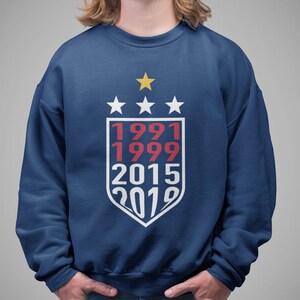 Puede incluir: Una sudadera azul marino con un dise&ntilde;o de escudo blanco que presenta los a&ntilde;os 1991, 1999, 2015 y 2019 en texto rojo y blanco. Hay cuatro estrellas blancas sobre el escudo y una estrella dorada sobre las estrellas.