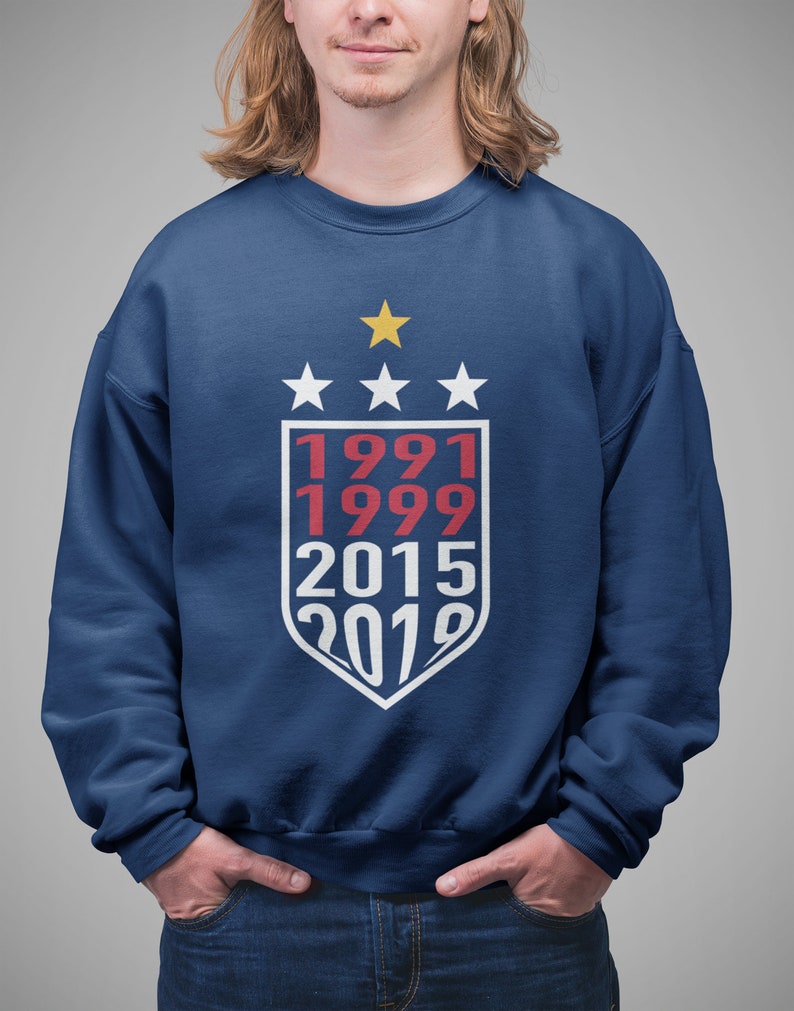 Puede incluir: Una sudadera azul marino con un dise&ntilde;o de escudo blanco que presenta los a&ntilde;os 1991, 1999, 2015 y 2019 en texto rojo y blanco. Hay cuatro estrellas blancas sobre el escudo y una estrella dorada sobre las estrellas.