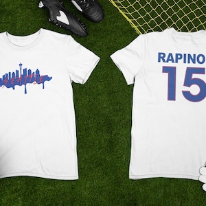 Puede incluir: Camiseta blanca con un gr&aacute;fico azul del horizonte de Seattle en la parte delantera y el texto "RAPINOE 15" en la parte posterior.