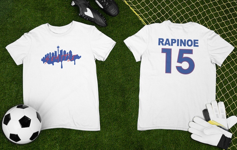 Puede incluir: Camiseta blanca con un gr&aacute;fico azul del horizonte de Seattle en la parte delantera y el texto "RAPINOE 15" en la parte posterior.