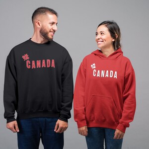 May include: Two people wearing sweatshirts with a maple leaf and the word "CANADA" printed on them. The person on the left is wearing a black sweatshirt with red text. The person on the right is wearing a red sweatshirt with white text.