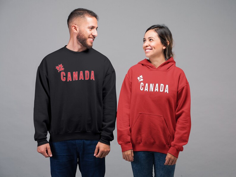 May include: Two people wearing sweatshirts with a maple leaf and the word "CANADA" printed on them. The person on the left is wearing a black sweatshirt with red text. The person on the right is wearing a red sweatshirt with white text.