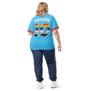 Op de afbeelding: Een lichtblauw T-shirt met het woord "Groom" en het nummer "10" gedrukt op de achterkant. Het nummer is omringd door een regenboogstreepontwerp.