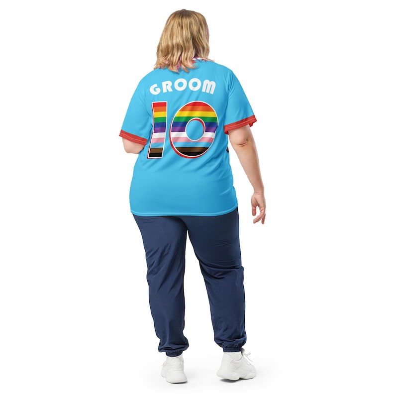 Op de afbeelding: Een lichtblauw T-shirt met het woord "Groom" en het nummer "10" gedrukt op de achterkant. Het nummer is omringd door een regenboogstreepontwerp.