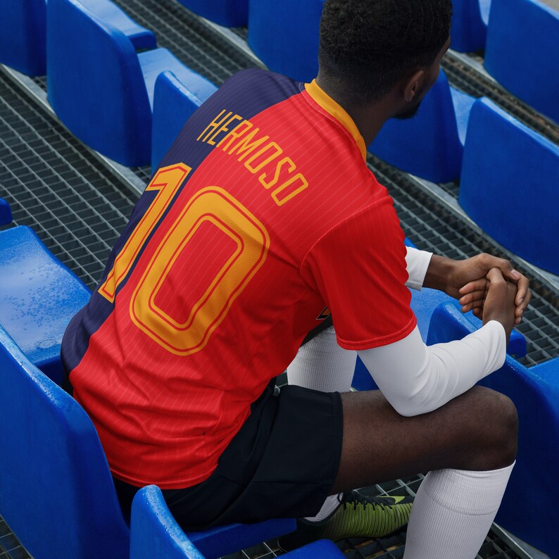 May include: A red and black soccer jersey with the number 10 and the name "HERMOSO" printed on the back. The jersey is worn by a person sitting on blue stadium bleachers.