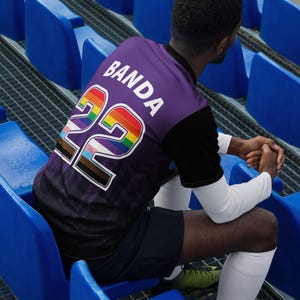 Op de afbeelding: Een paars voetbaltenue met een regenboognummer 22 en het woord "Banda" op de rug. Het tenue wordt gedragen door een persoon die op blauwe stadionbanken zit.