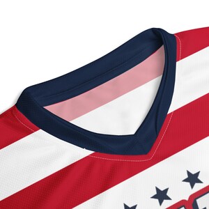 May include: A red, white, and blue striped jersey with a navy blue v-neck collar. The jersey has a pink stripe under the collar.