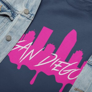 May include: A navy blue t-shirt featuring a pink silhouette of the San Diego skyline with the text "San Diego" in white. The skyline is dripping with pink paint.