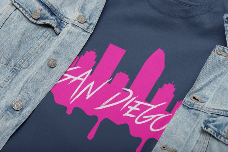 May include: A navy blue t-shirt featuring a pink silhouette of the San Diego skyline with the text "San Diego" in white. The skyline is dripping with pink paint.