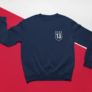 K&ouml;nnte beinhalten: Marineblauer Sweatshirt mit einem wei&szlig;en gestickten Schild mit der Nummer 13 und f&uuml;nf Sternen.