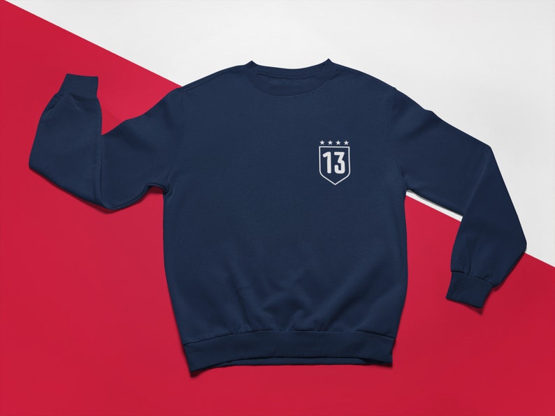K&ouml;nnte beinhalten: Marineblauer Sweatshirt mit einem wei&szlig;en gestickten Schild mit der Nummer 13 und f&uuml;nf Sternen.