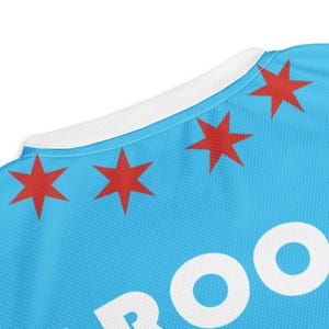 Op de afbeelding: Een lichtblauw T-shirt met een witte kraag en witte tekst die "Chicago" zegt met een witte omtrek. Het shirt heeft vier rode sterren op de bovenste helft van het shirt.