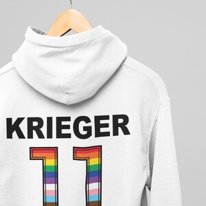 Puede incluir: Sudadera con capucha blanca con la palabra "KRIEGER" impresa en negro y el n&uacute;mero "11" en colores arco&iacute;ris. El n&uacute;mero est&aacute; contorneado en negro.