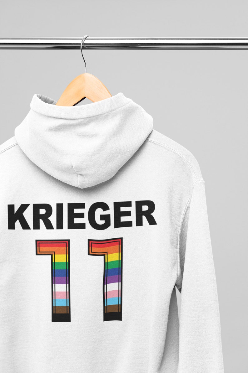 Puede incluir: Sudadera con capucha blanca con la palabra "KRIEGER" impresa en negro y el n&uacute;mero "11" en colores arco&iacute;ris. El n&uacute;mero est&aacute; contorneado en negro.