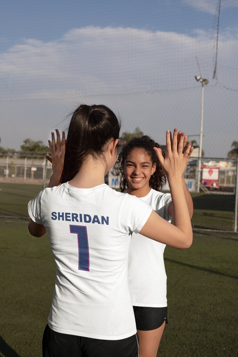 May include: Two young women wearing white soccer jerseys with the text "SHERIDAN" and the number "1" on the back. They are high-fiving each other on a soccer field.
