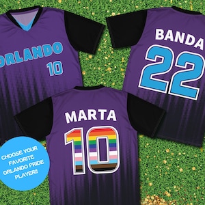 Op de afbeelding: Drie paarse voetbalshirts met zwarte mouwen. De shirts hebben verschillende namen en nummers op de rug. Het eerste shirt heeft "ORLANDO 10" op de rug. Het tweede shirt heeft "BANDA 22" op de rug. Het derde shirt heeft "MARTHA 10" op de rug met een regenboognummer.