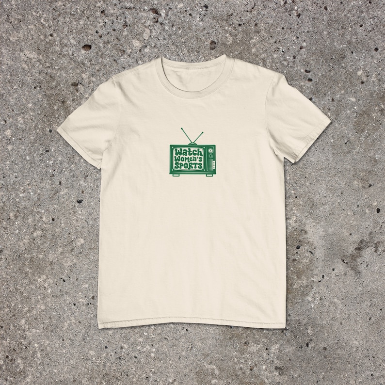 May include: A cream colored t-shirt with a green retro television graphic that says "Watch Women's Sports" on the screen.