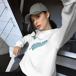 May include: White sweatshirt with a teal green fern and the text "NEW ZEALAND" printed on the front. The model is wearing a grey baseball cap.