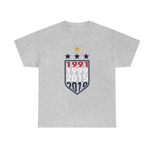 Puede incluir: Una camiseta gris con un dise&ntilde;o de escudo azul marino y rojo que presenta los a&ntilde;os 1991, 1999, 2015 y 2019. Hay tres estrellas blancas y una estrella amarilla sobre el escudo.