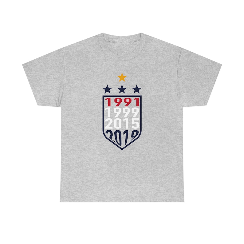 Puede incluir: Una camiseta gris con un dise&ntilde;o de escudo azul marino y rojo que presenta los a&ntilde;os 1991, 1999, 2015 y 2019. Hay tres estrellas blancas y una estrella amarilla sobre el escudo.