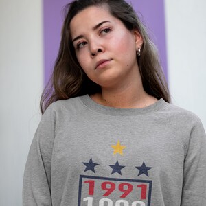 Puede incluir: Una sudadera gris con un gr&aacute;fico de estilo vintage que presenta tres estrellas, los a&ntilde;os 1991, 1999 y 2015, y una estrella amarilla.