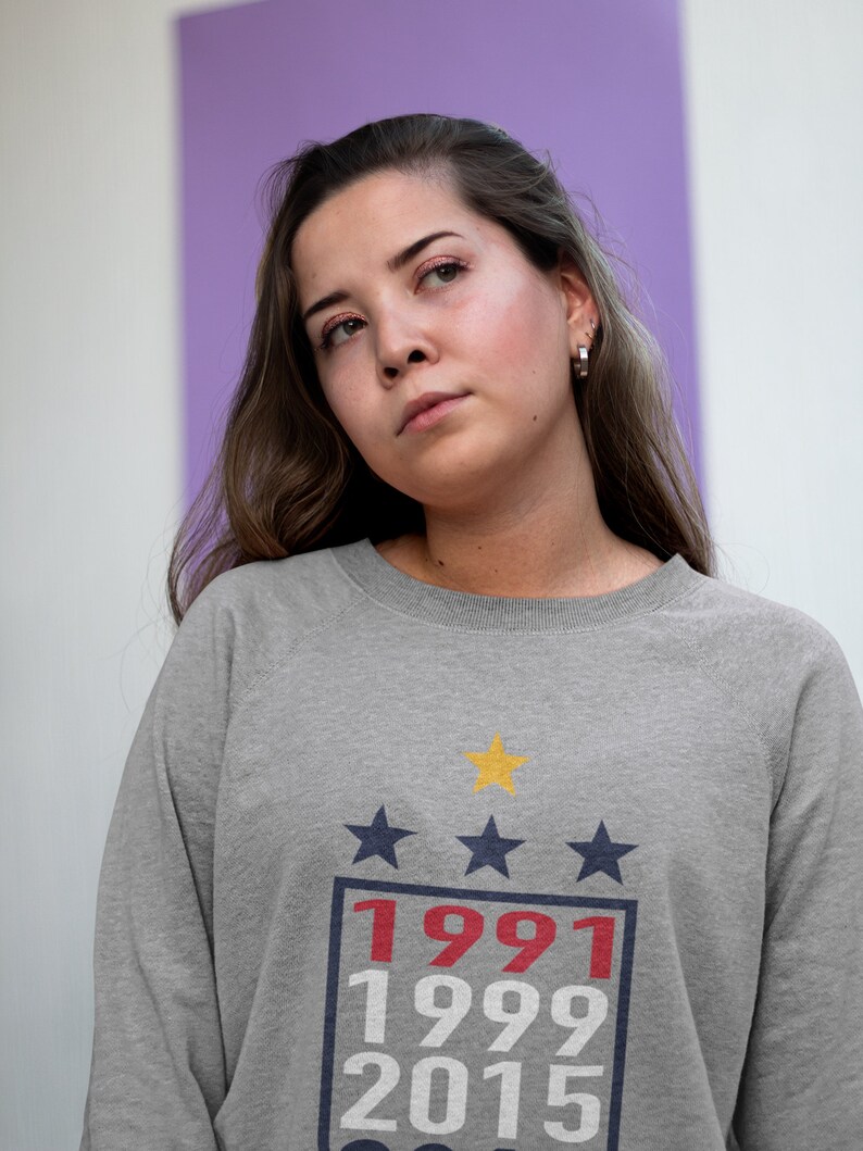 Puede incluir: Una sudadera gris con un gr&aacute;fico de estilo vintage que presenta tres estrellas, los a&ntilde;os 1991, 1999 y 2015, y una estrella amarilla.