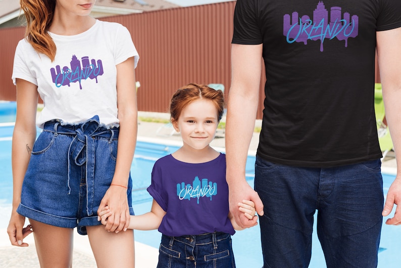 May include: A family of three wearing matching t-shirts with a graphic of the Orlando skyline and the word "Orlando" in a dripping paint style. The shirts are white, black, and purple.