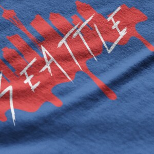 Puede incluir: Un dise&ntilde;o gr&aacute;fico rojo y blanco sobre una camiseta azul con la palabra "SEATTLE" y una silueta de horizonte.
