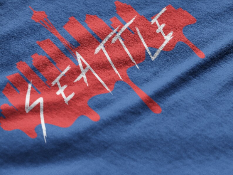 Puede incluir: Un dise&ntilde;o gr&aacute;fico rojo y blanco sobre una camiseta azul con la palabra "SEATTLE" y una silueta de horizonte.