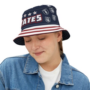 May include: A blue and red bucket hat with a white star pattern and the word "STATES" in white letters. The hat also has red and white stripes around the brim and features numbers and names on the front.