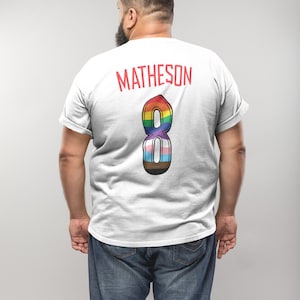 May include: White t-shirt with the name "MATHESON" in red block letters across the upper back. Below the name is a large number "8" in a rainbow design, incorporating various pride flag colours. The person is wearing blue jeans.