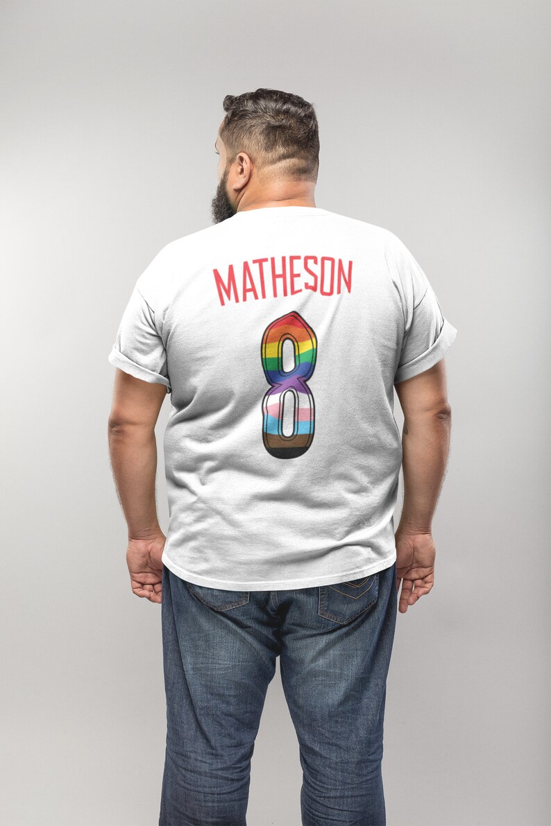 May include: White t-shirt with the name "MATHESON" in red block letters across the upper back. Below the name is a large number "8" in a rainbow design, incorporating various pride flag colours. The person is wearing blue jeans.