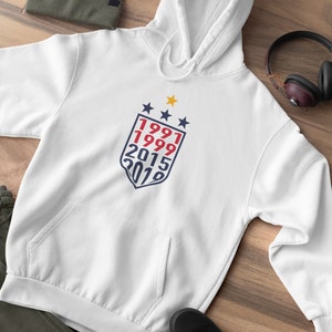May include: White hooded sweatshirt with a blue and red shield graphic featuring the years 1991, 1999, 2015, and 2019. There are three gold stars above the shield.