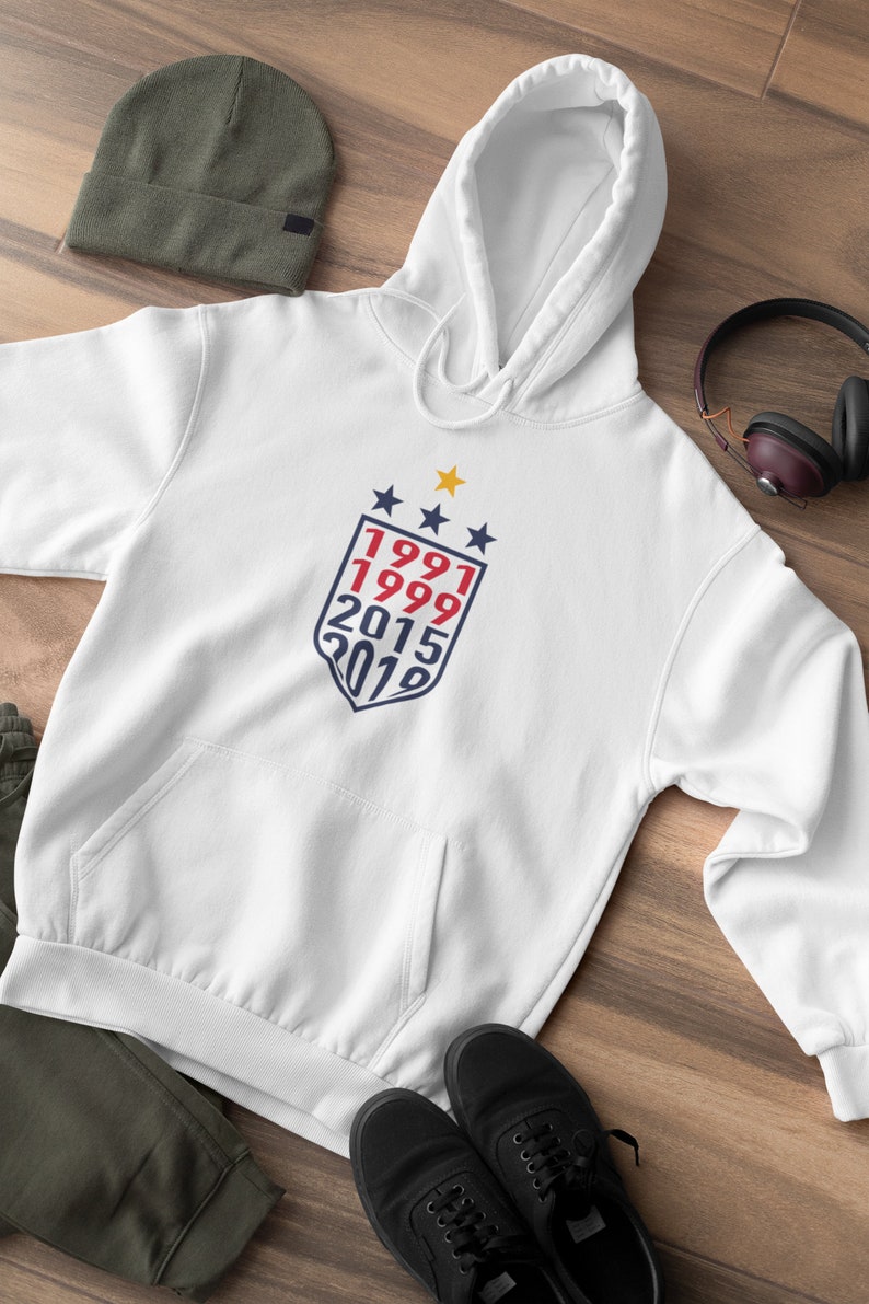 May include: White hooded sweatshirt with a blue and red shield graphic featuring the years 1991, 1999, 2015, and 2019. There are three gold stars above the shield.